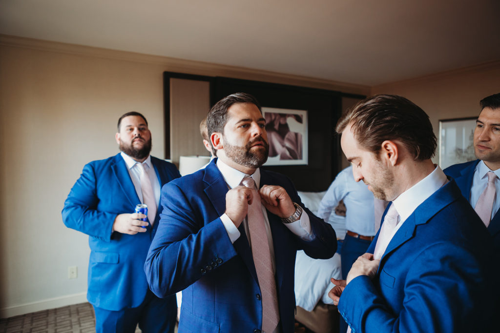 a groom fixes his tie with his groomsmen in a hotel room