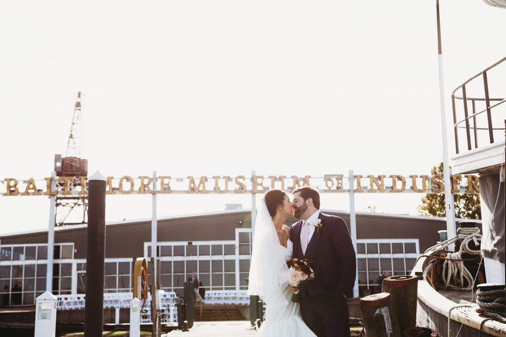 a bride and groom stand in the sun in front of the baltimore museum of industry